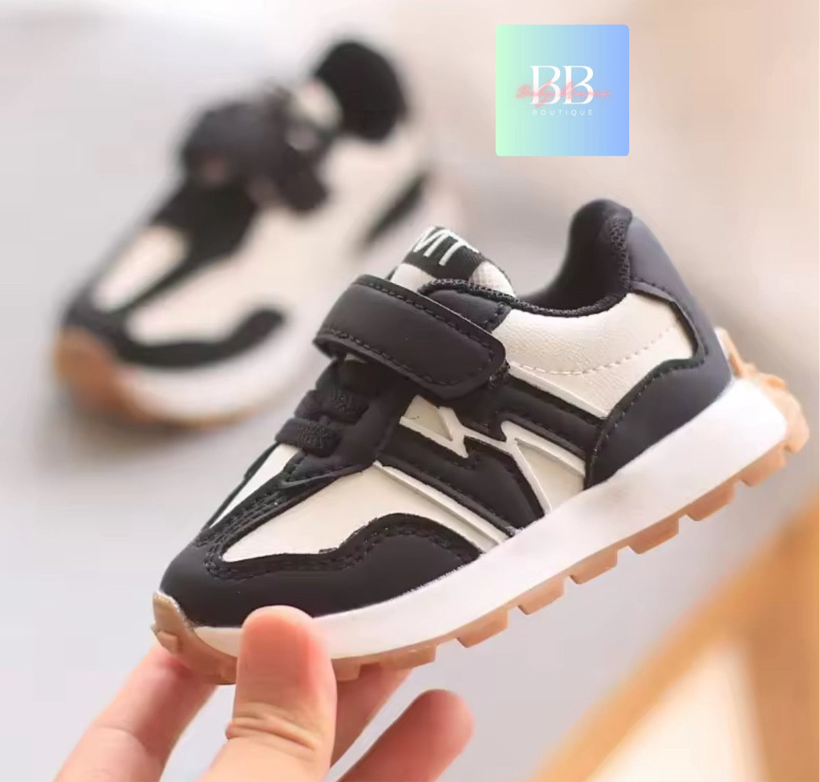 Black and white children's sneakers with velcro straps, held by a hand against a blurred background.