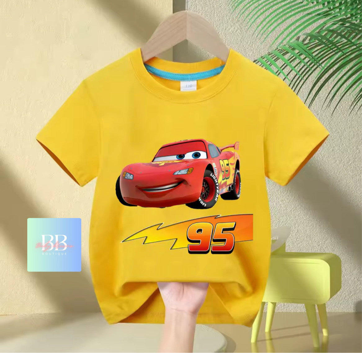 Yellow t-shirt with a red car graphic and number 95 on a hanger against a beige wall.