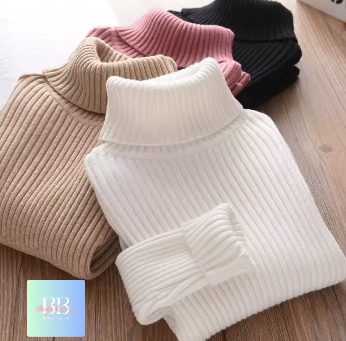 Three knitted turtleneck sweaters in beige, pink, and black on a wooden surface.