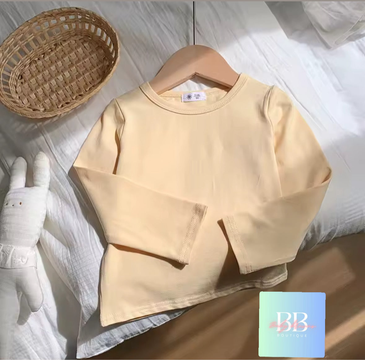 Beige long-sleeve shirt on a hanger with a woven basket and plush toy on a light background