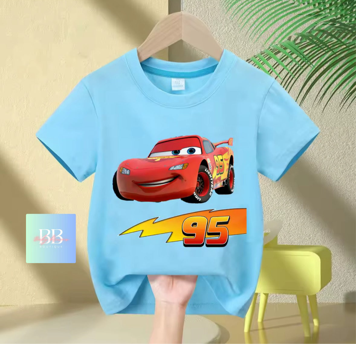 Light blue t-shirt with a red car graphic and number 95 on a hanger against a beige wall.