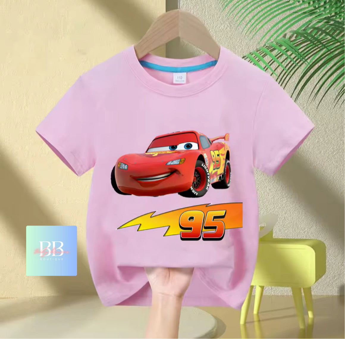 Pink t-shirt with a cartoon car design on a hanger against a light-colored wall.