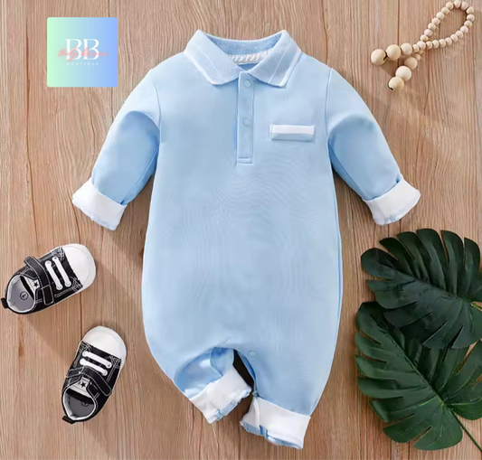 Blue baby onesie with white trim on a wooden surface with baby shoes and a leaf.