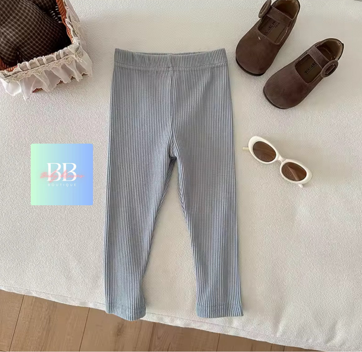 Gray ribbed leggings with brown shoes and sunglasses on a white surface, featuring a brand logo in the corner.