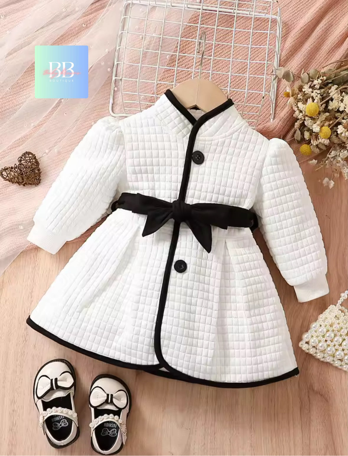 White quilted dress with black buttons and belt on a wooden surface, accompanied by black shoes.