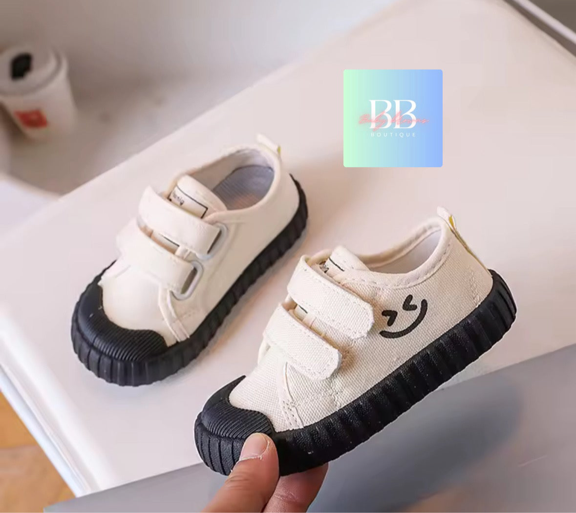 Pair of white children's shoes with black soles held by a hand, on a light background.