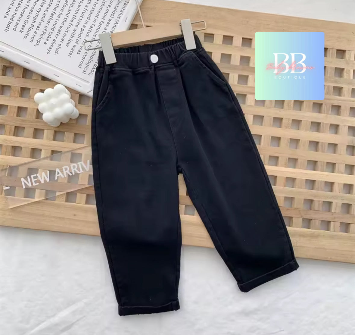 Black pants hanging on a wooden rack with a 'NEW ARRIVAL' sign and a BB card in the background.