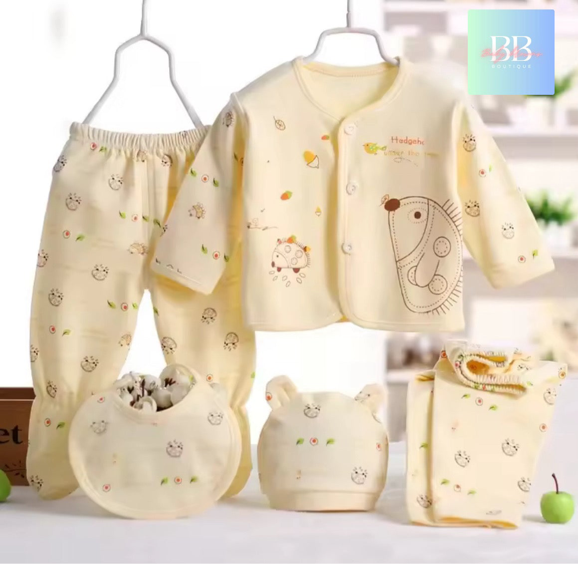 Baby clothing set with a yellow outfit, pants, bib, and hat on hangers with a blurred background.