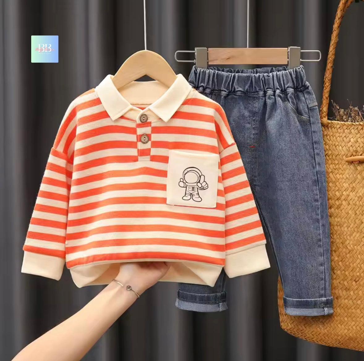 Children's striped shirt with a pocket and jeans on hangers against a dark curtain background.