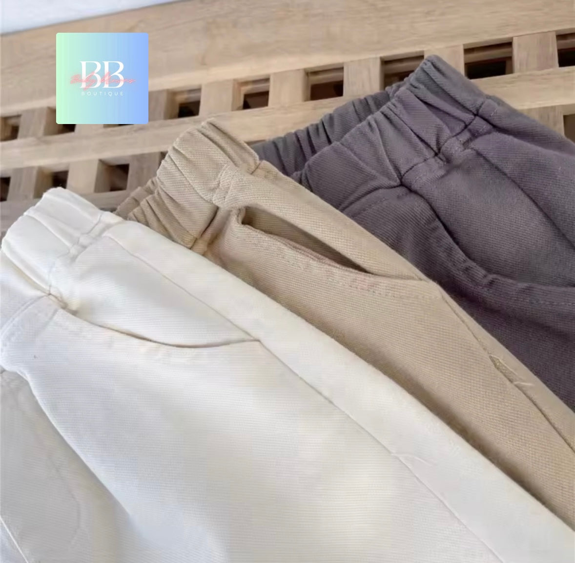 Three pairs of pants in white, beige, and gray on a wooden surface with a brand logo in the corner.