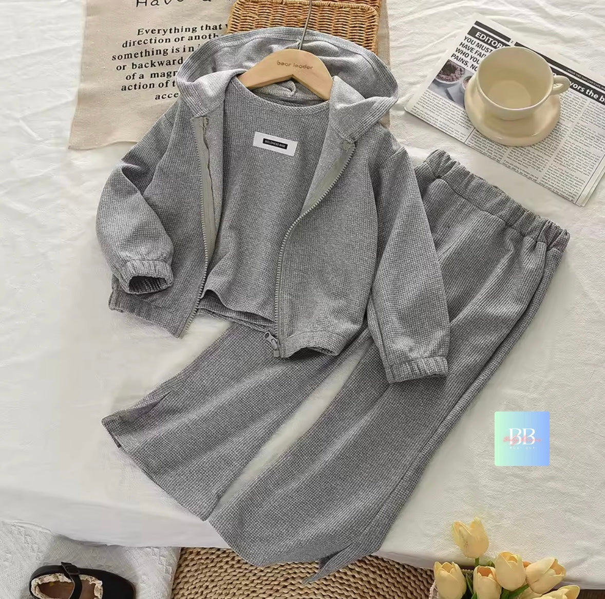 Gray children's outfit with a jacket and pants on a bed with a newspaper and cup.