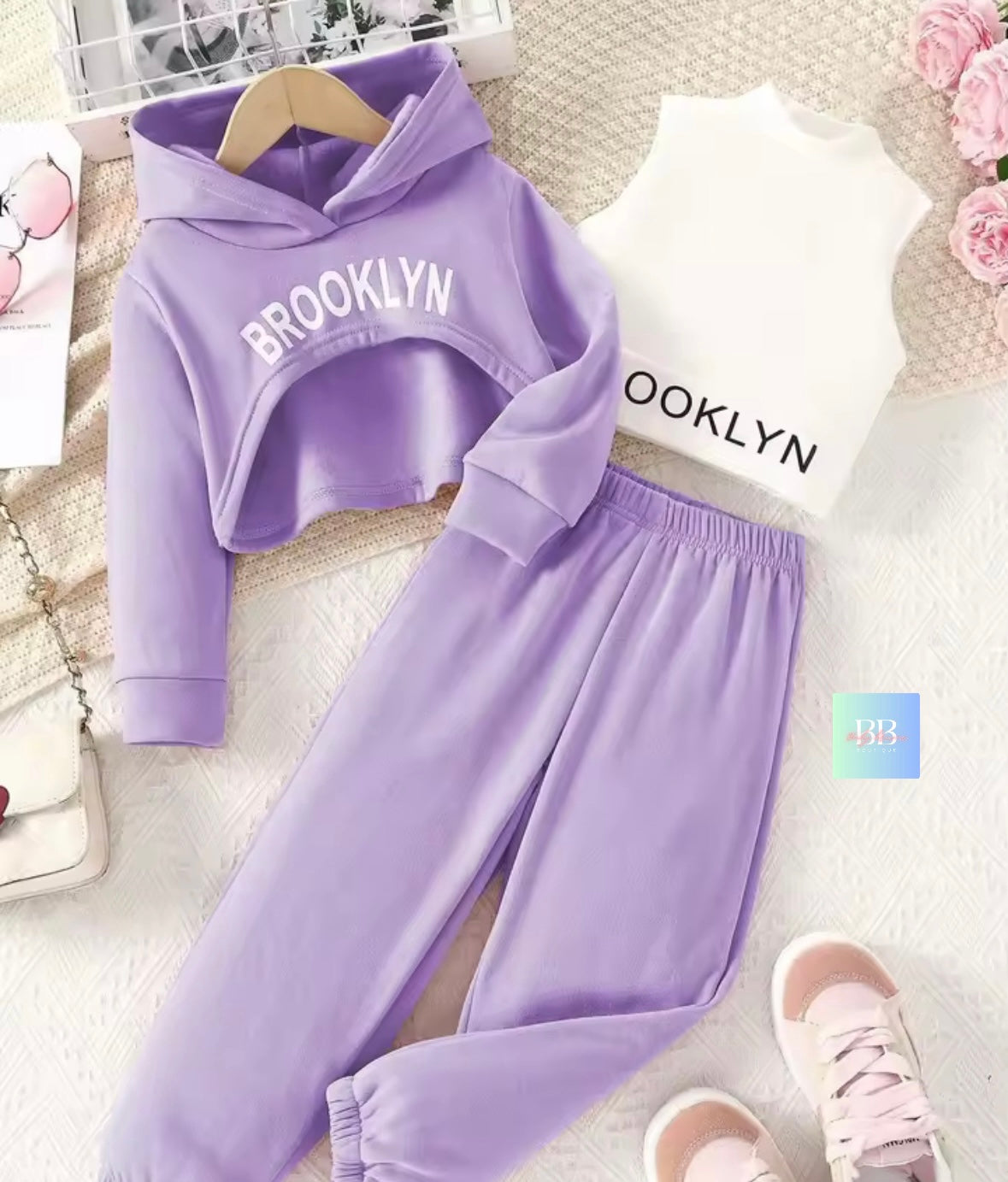 Purple 'Brooklyn' hoodie and pants set on a textured surface with pink shoes and flowers.