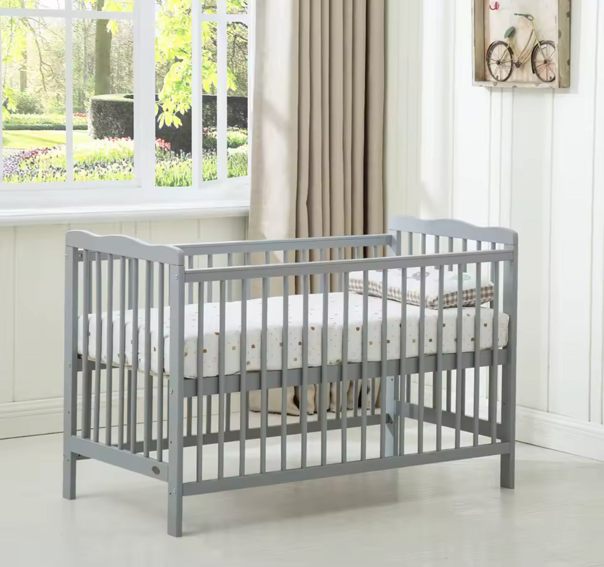 Gray crib with white mattress in a room with a window and curtains.