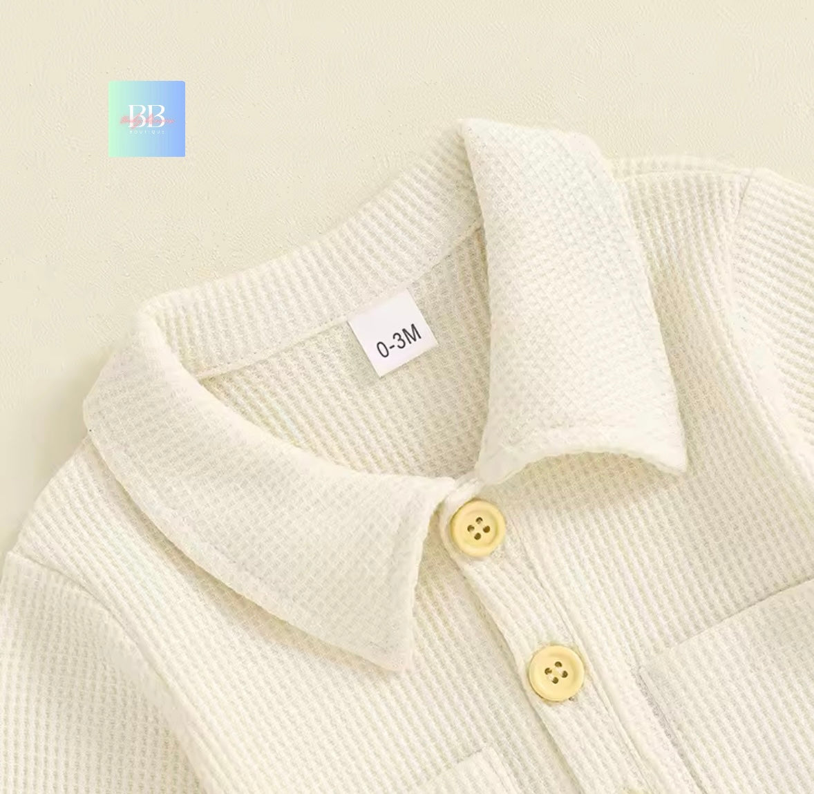 Close-up of a baby's white textured outfit with gold buttons on a beige background