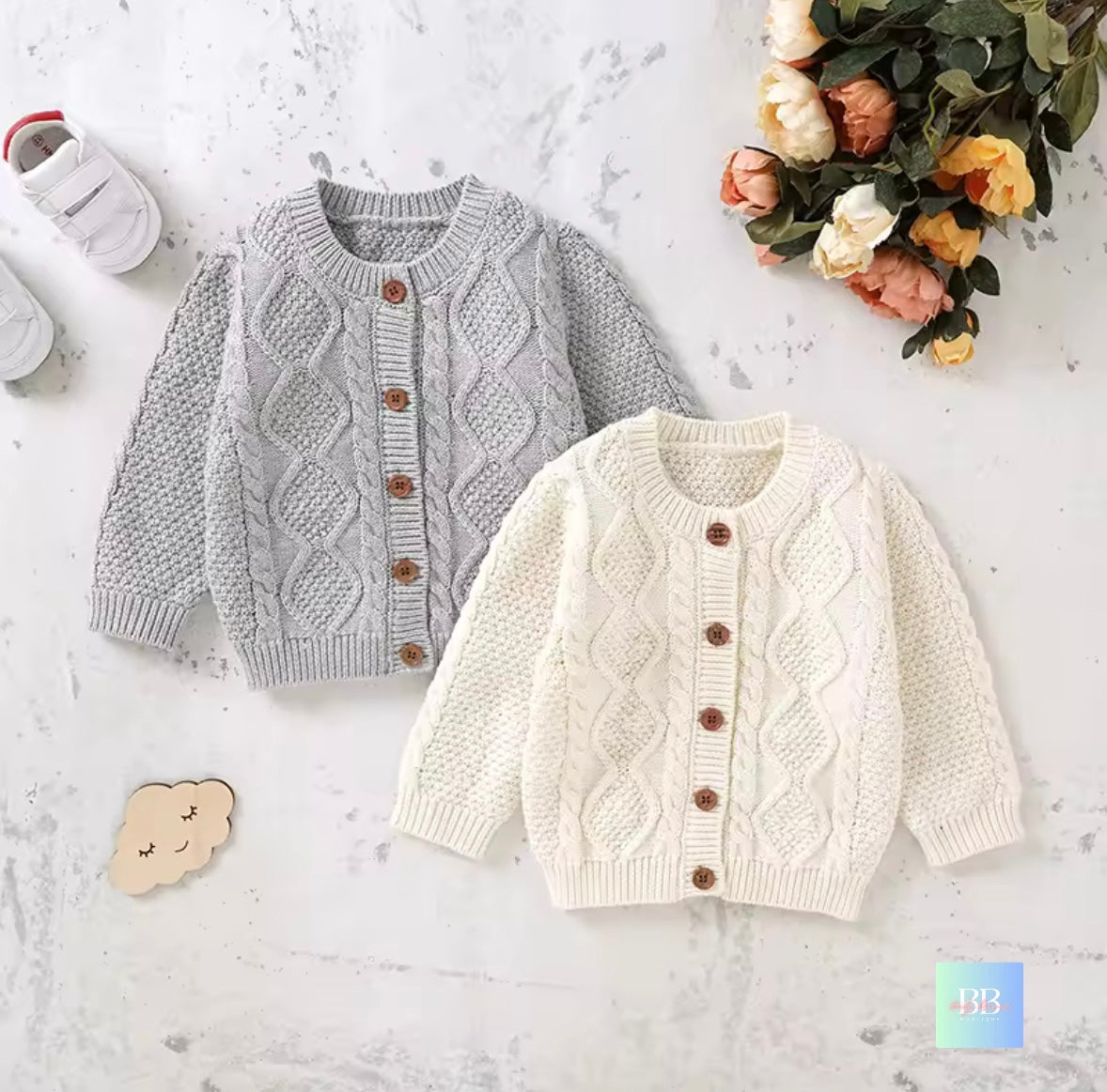 Two cable knit cardigans, one gray and one white, on a light surface with flowers and shoes.