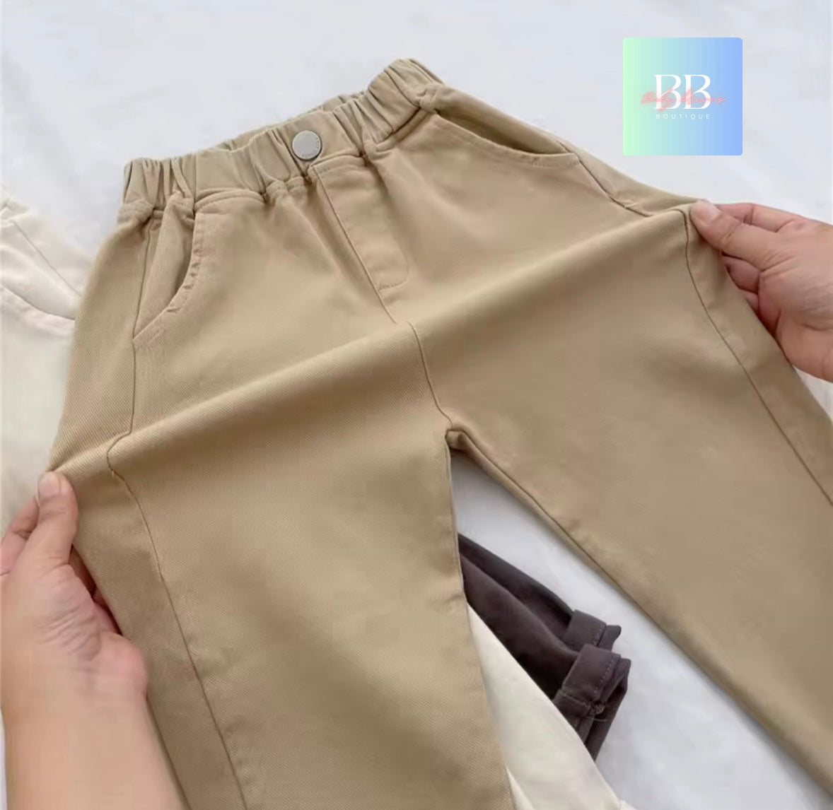 Beige pants being held up on a white background with a brand logo in the corner.
