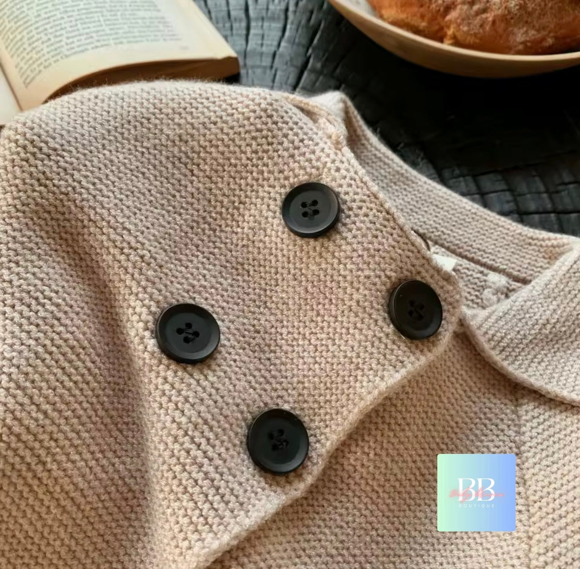 Close-up of a beige knitted garment with black buttons, next to an open book and bread on a wooden surface.