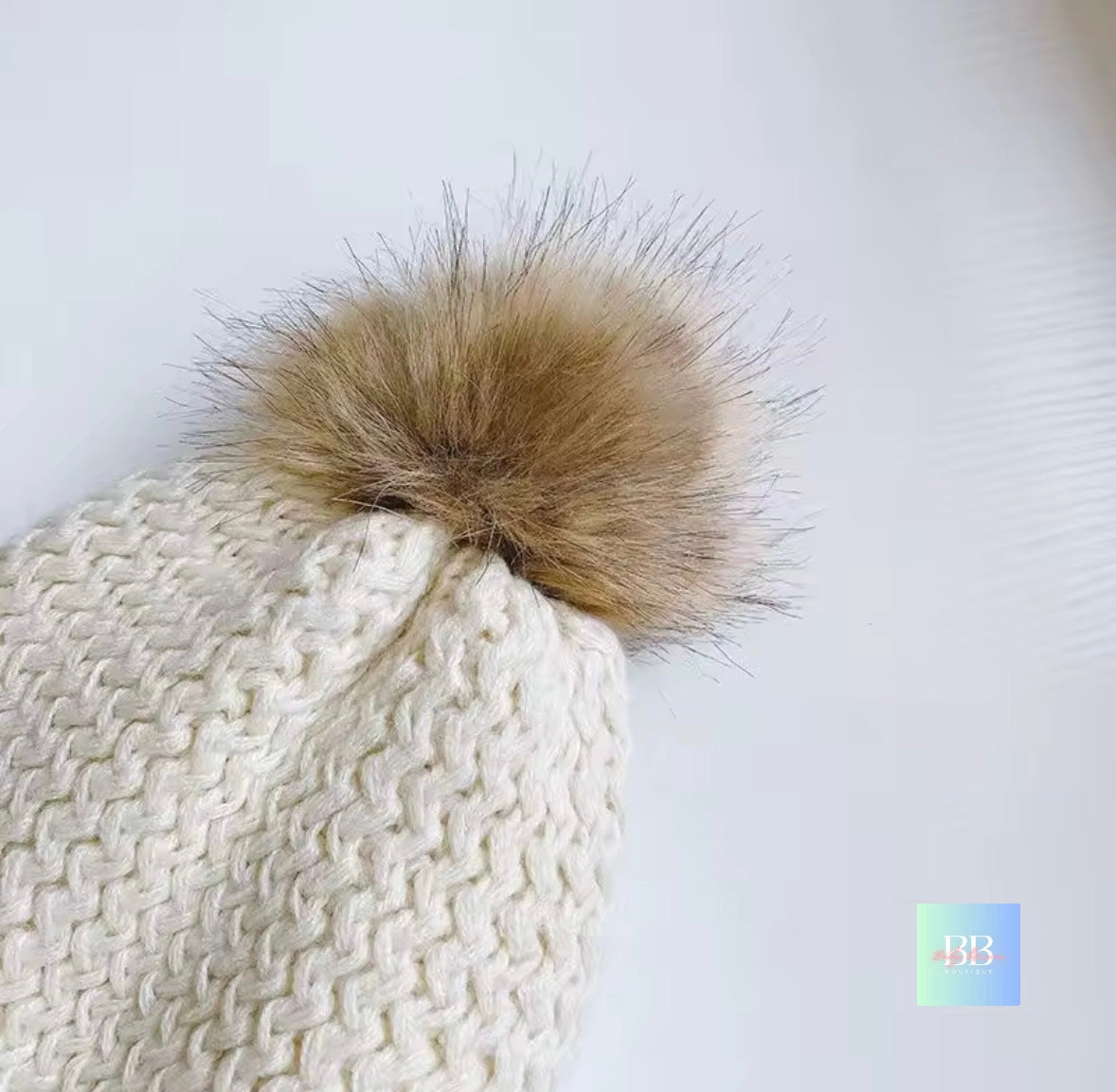 White knitted beanie with a large brown fur pom-pom on a light gray background