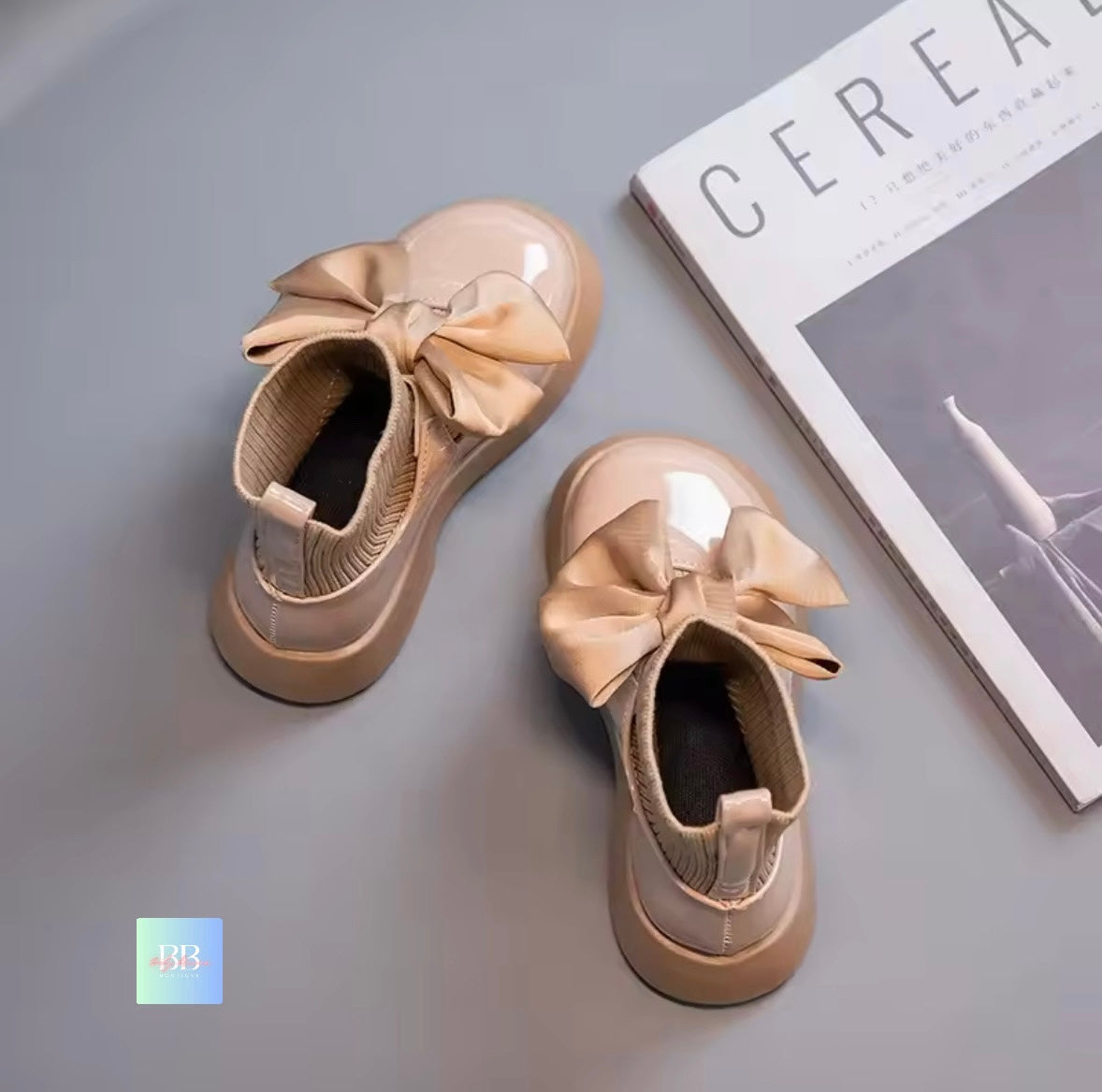 Pair of beige shoes with large bows on a gray surface next to a magazine.