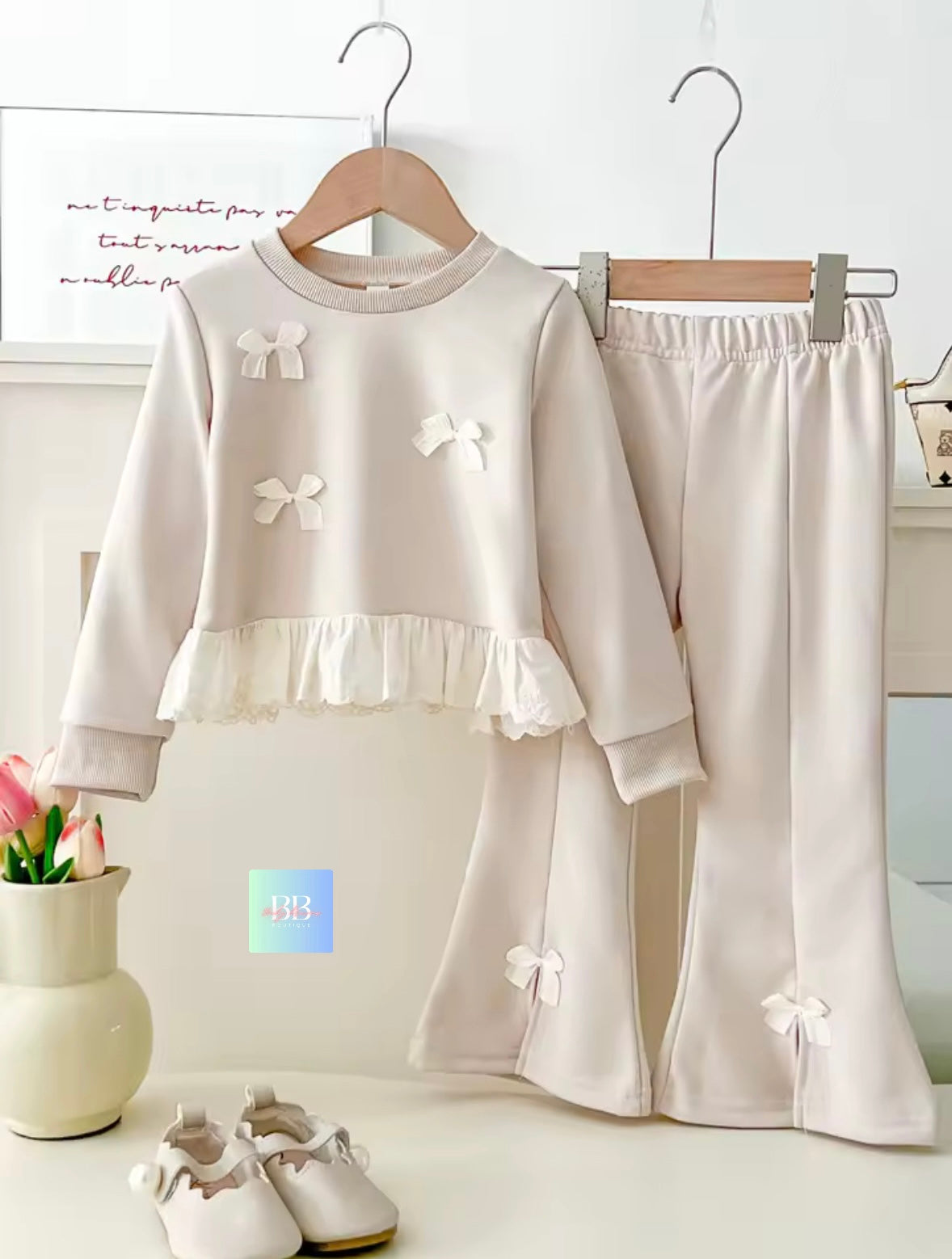 Children's outfit with white top and pants featuring bow details, displayed on hangers against a neutral background.