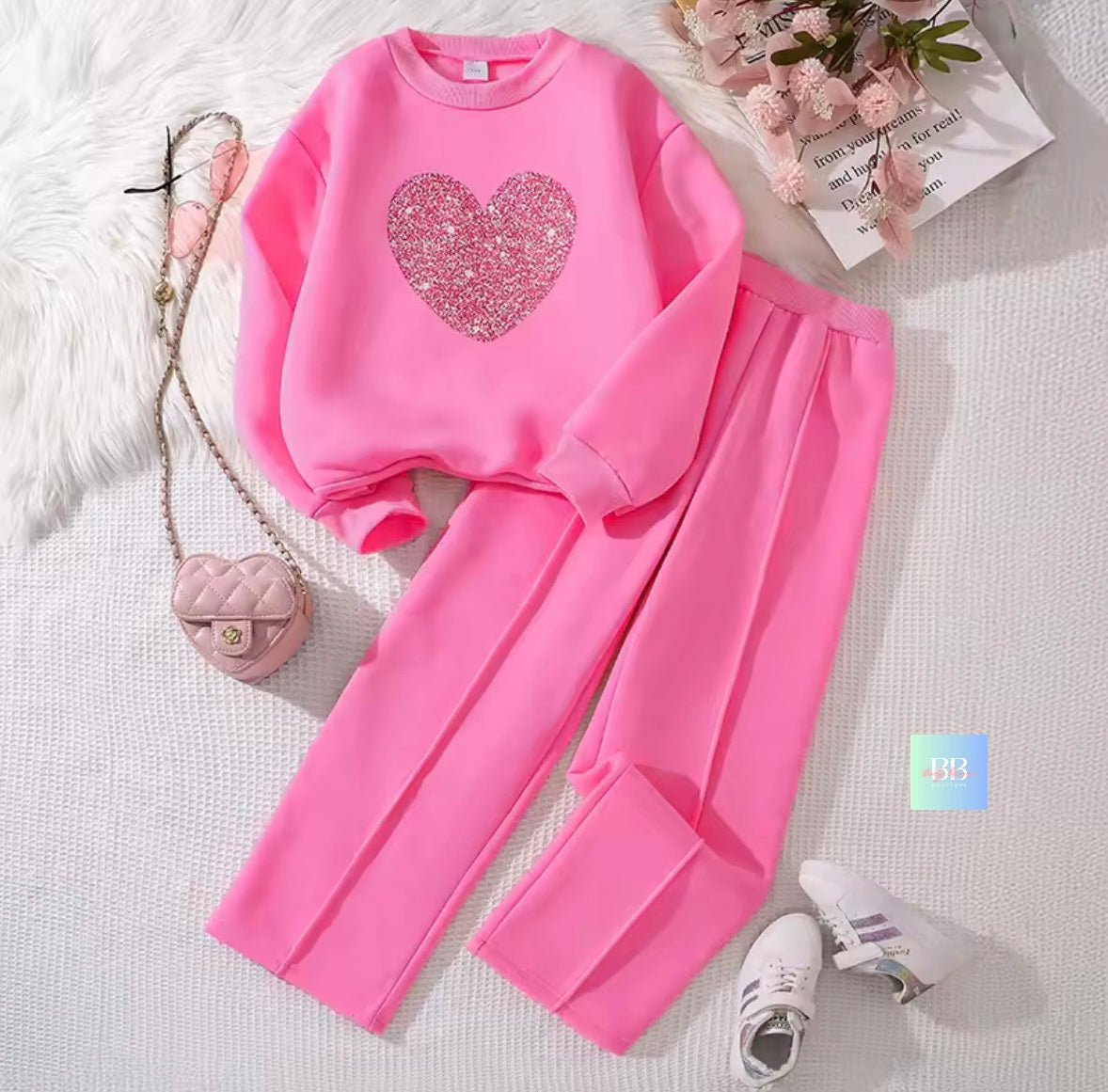 Pink sweatshirt with heart design and matching pants on a white surface with a small bag and shoes.