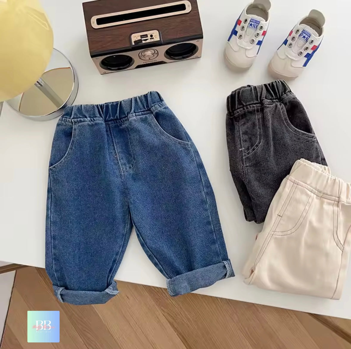 Three pairs of pants (blue, black, beige) on a white surface with a small wooden radio and baby shoes.