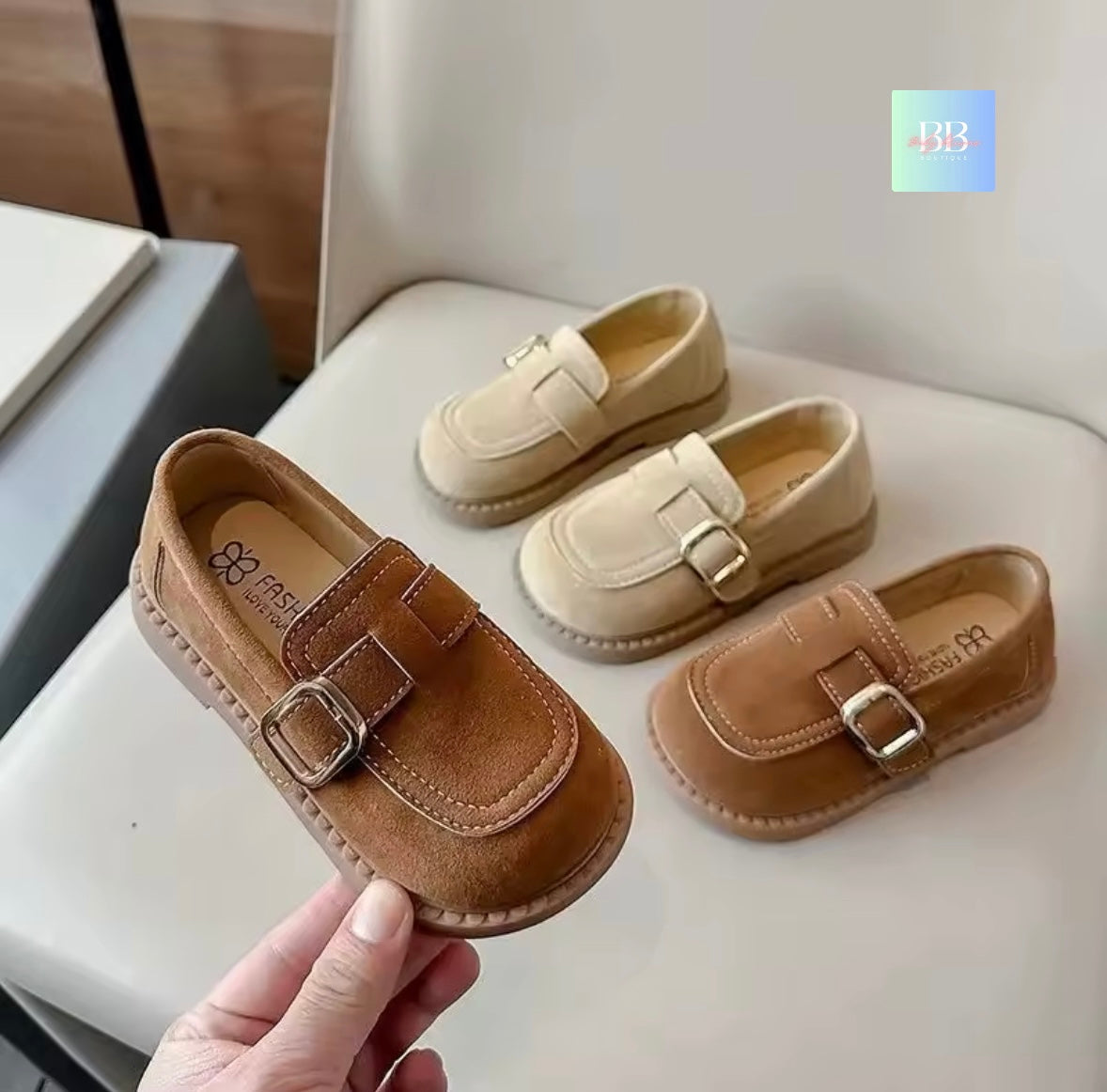 Pair of brown and beige children's loafers on a white surface with a hand holding one shoe.