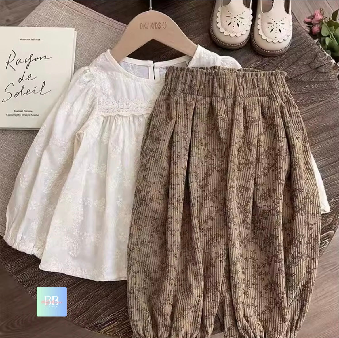 White blouse and brown patterned pants on a wooden surface with shoes in the background.