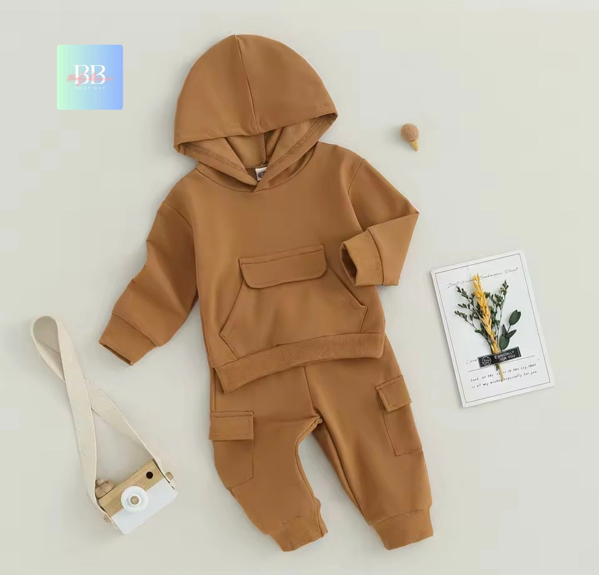 Brown hoodie and pants set on a light gray background with a camera and plant decoration.