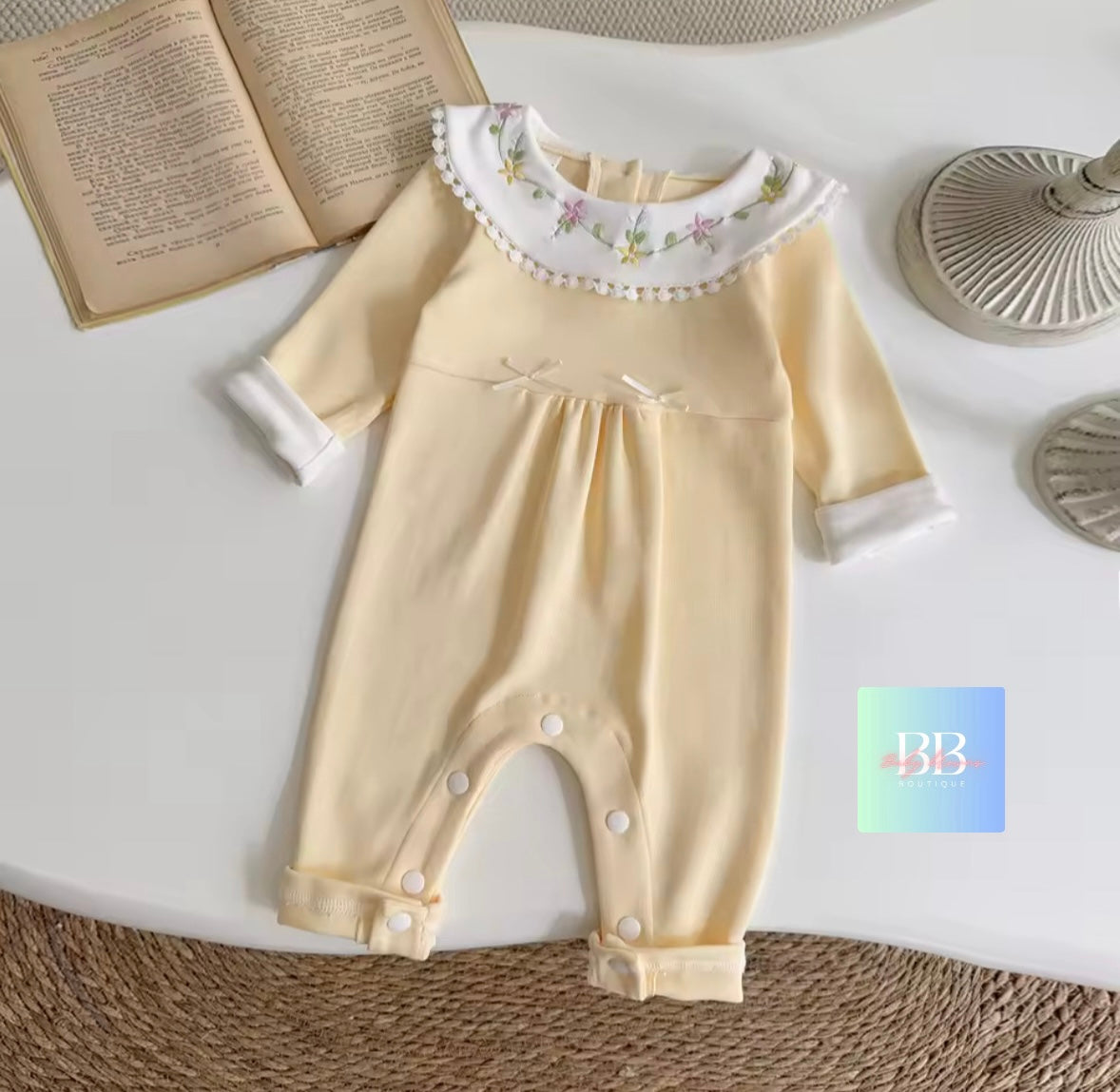 Baby romper with floral collar on a white surface, surrounded by books and decorative items.
