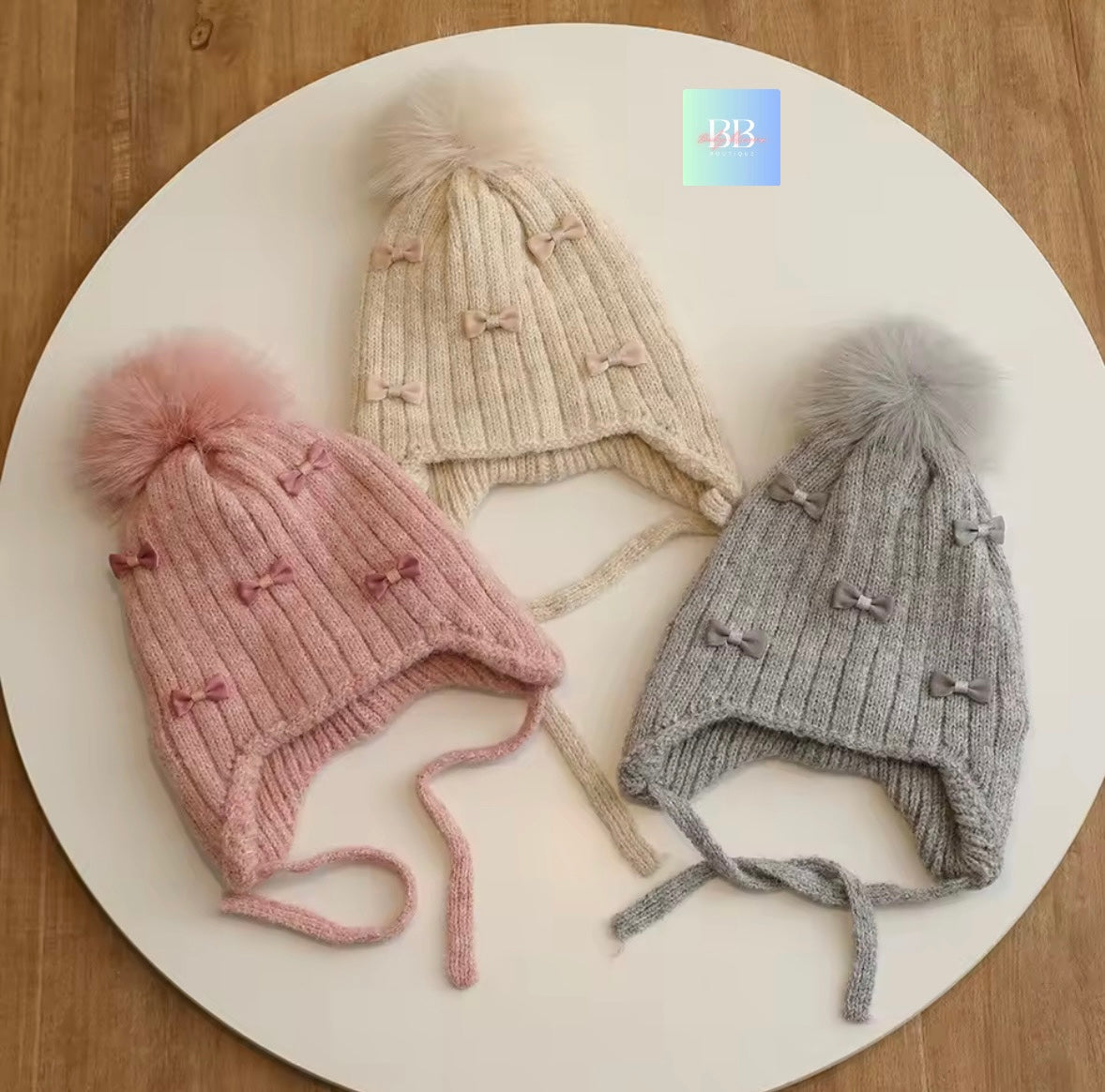 Three knitted hats with pom-poms in pink, beige, and gray on a white plate.