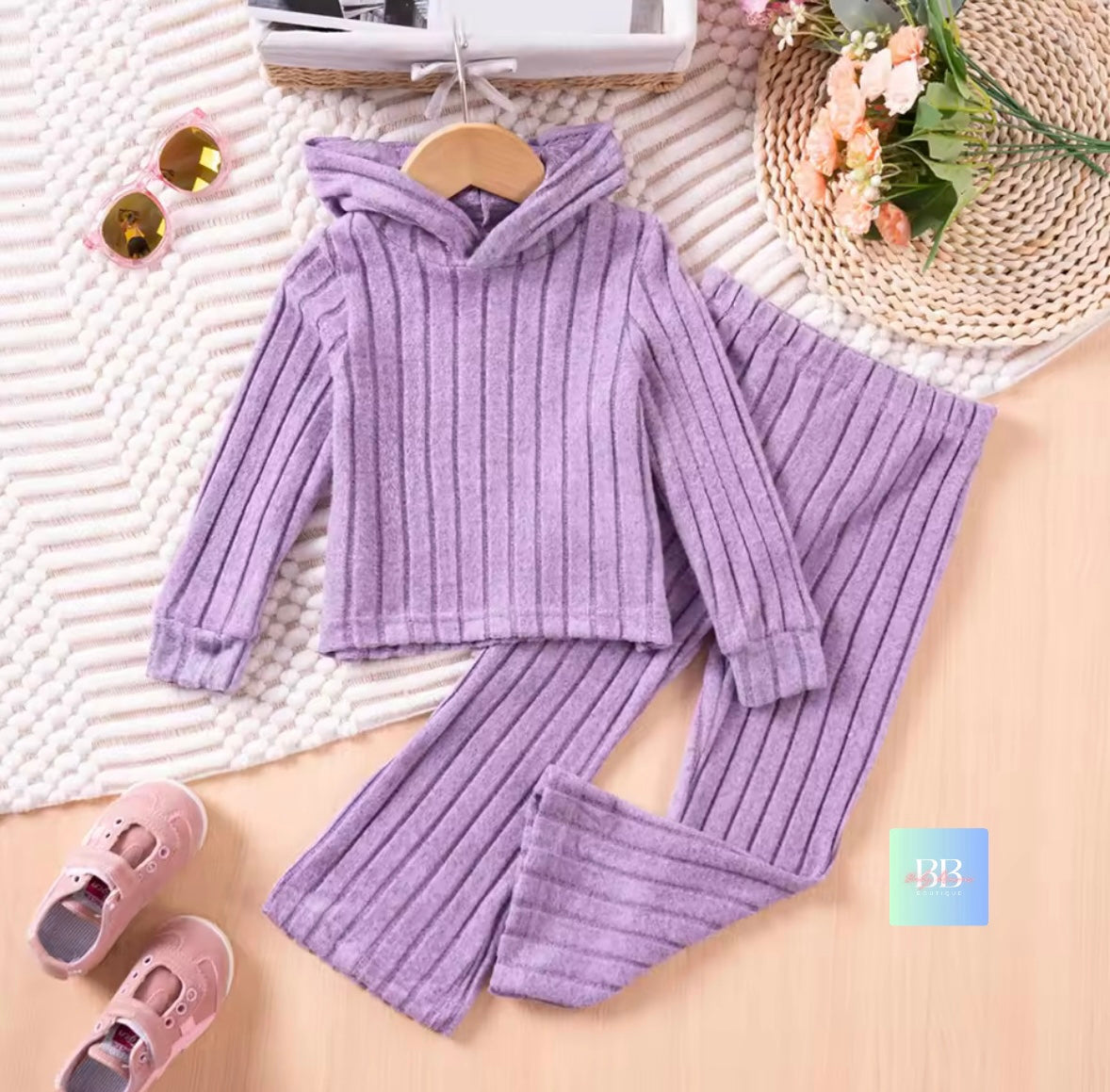 Purple ribbed hoodie and pants set on a textured surface with shoes and flowers.