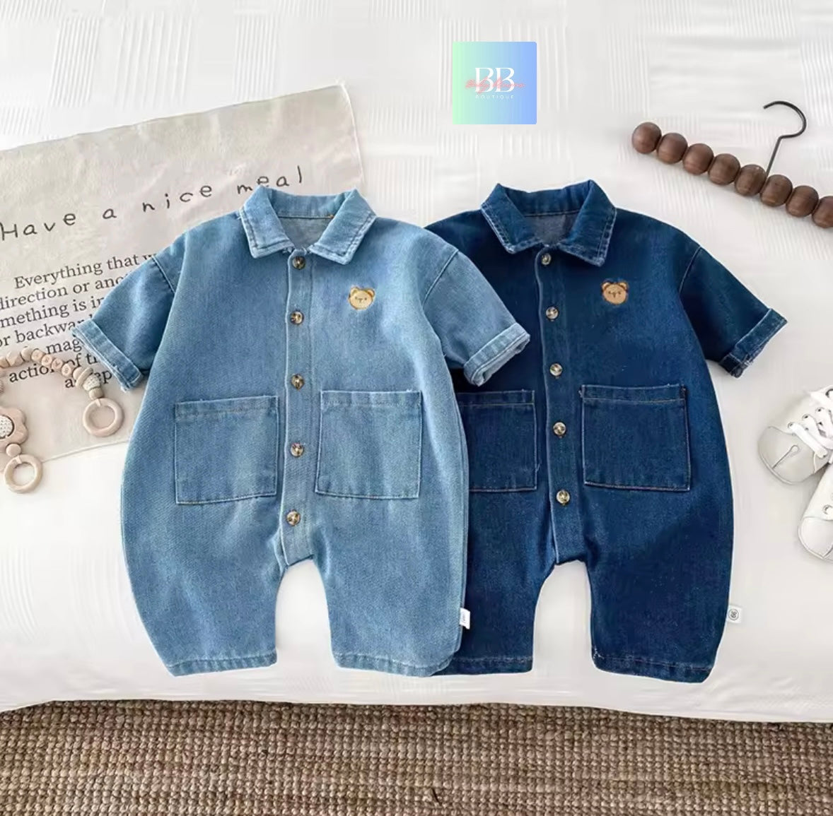 Two denim baby rompers on a textured surface with a magazine and hanger in the background.