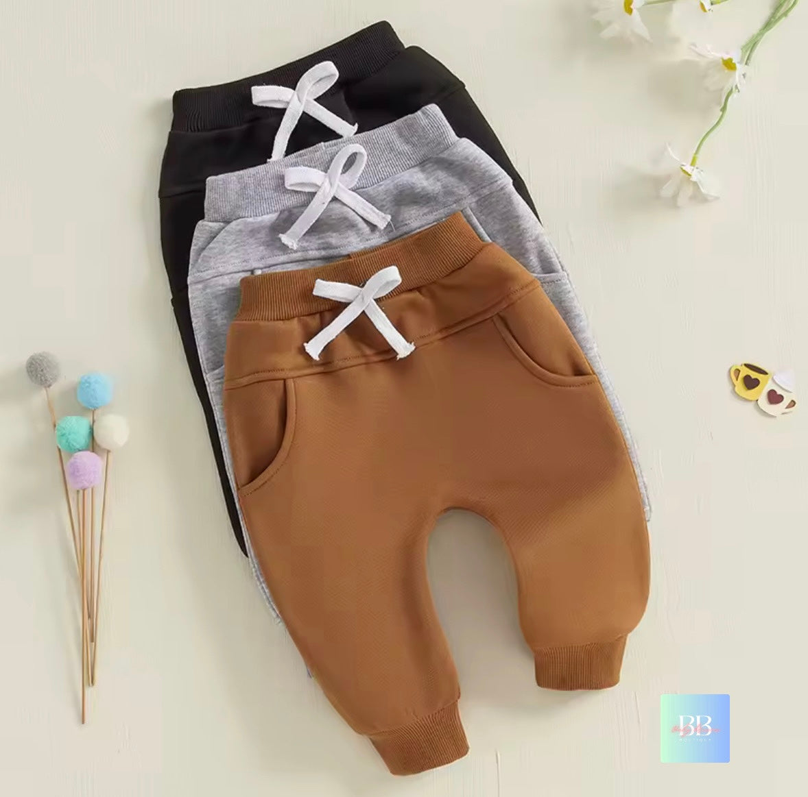 Three pairs of children's jogger pants in black, gray, and brown on a light background.