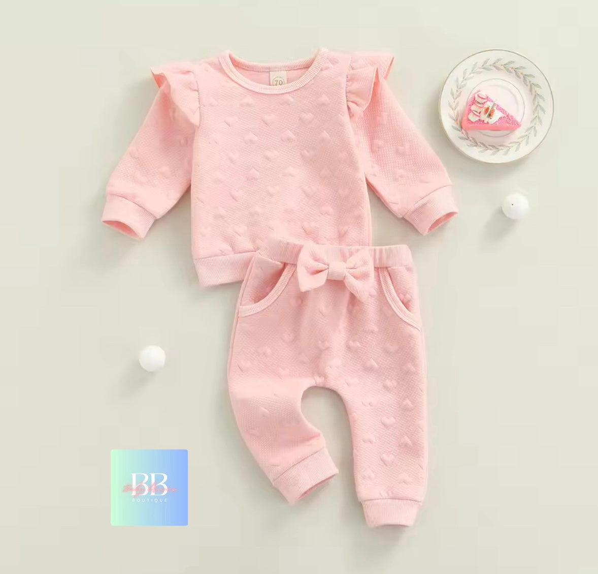 Pink baby outfit with ruffled sleeves and bow details on a light gray background.