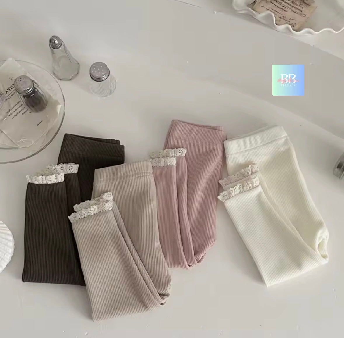 Four pairs of ribbed leggings with lace trim in black, gray, pink, and white on a white surface.