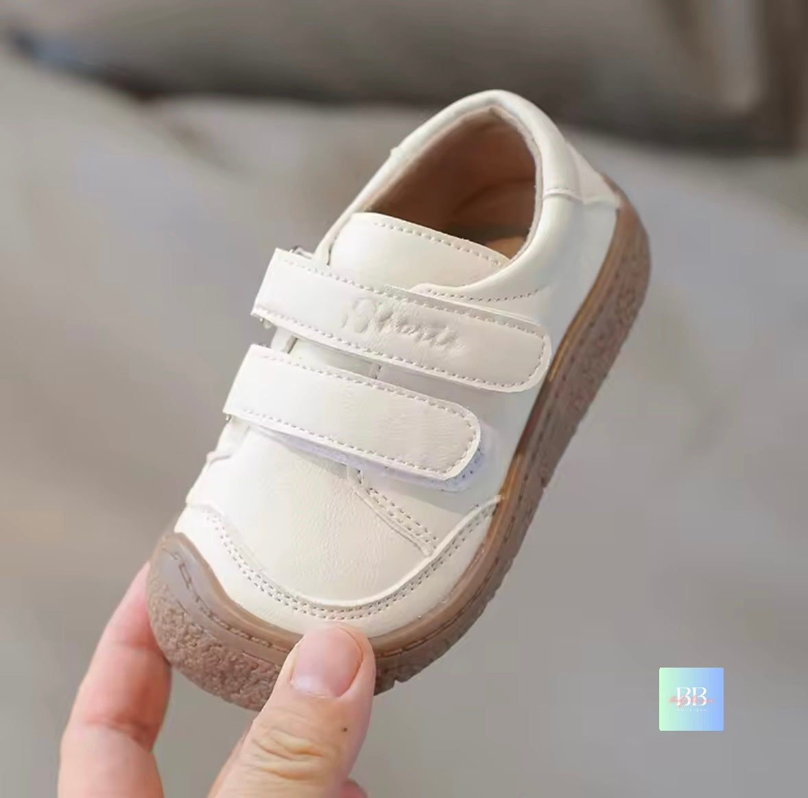 White children's shoe with brown sole held by a hand against a neutral background