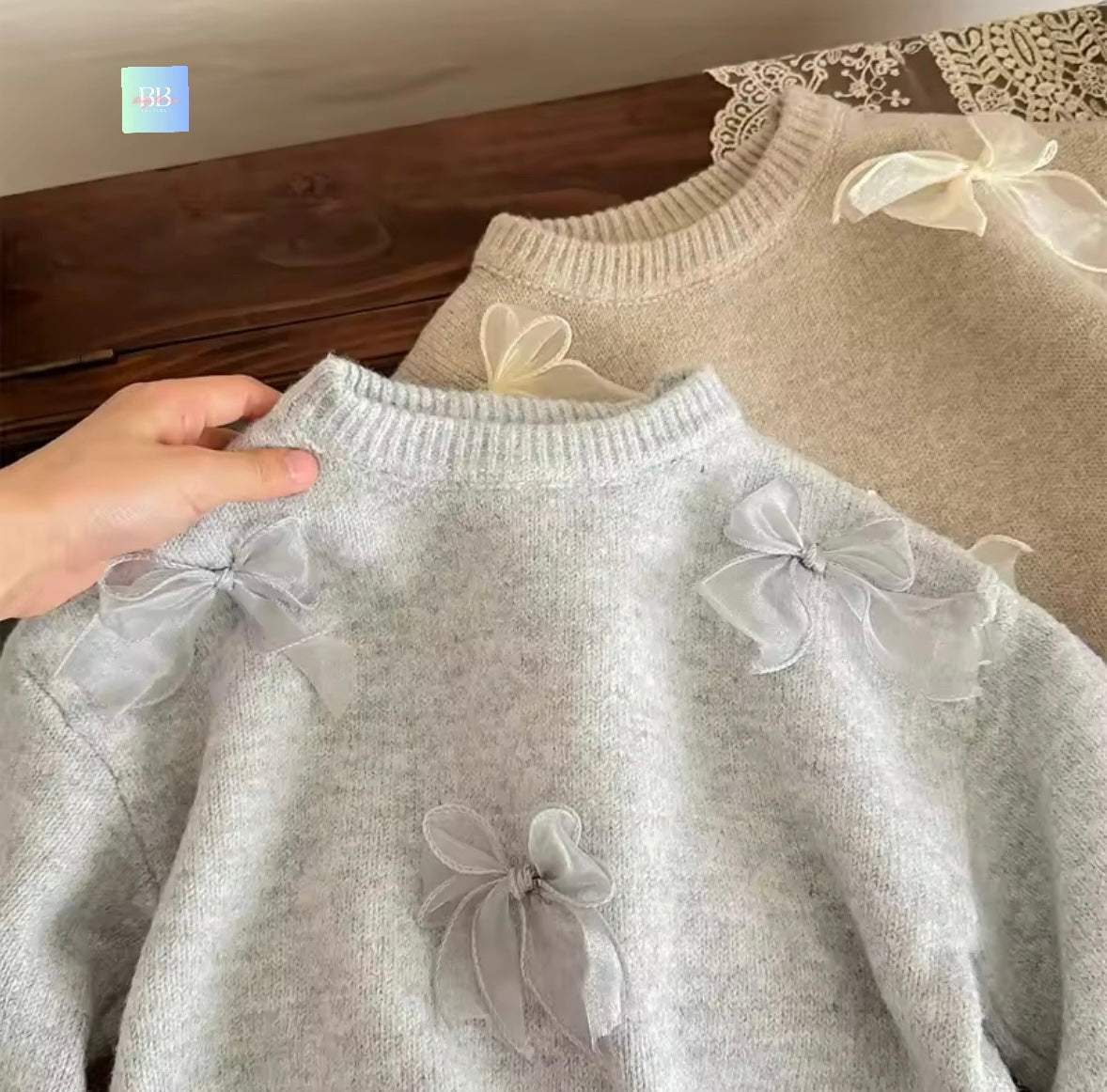 Gray sweater with sheer bow details held by a hand against a wooden surface.