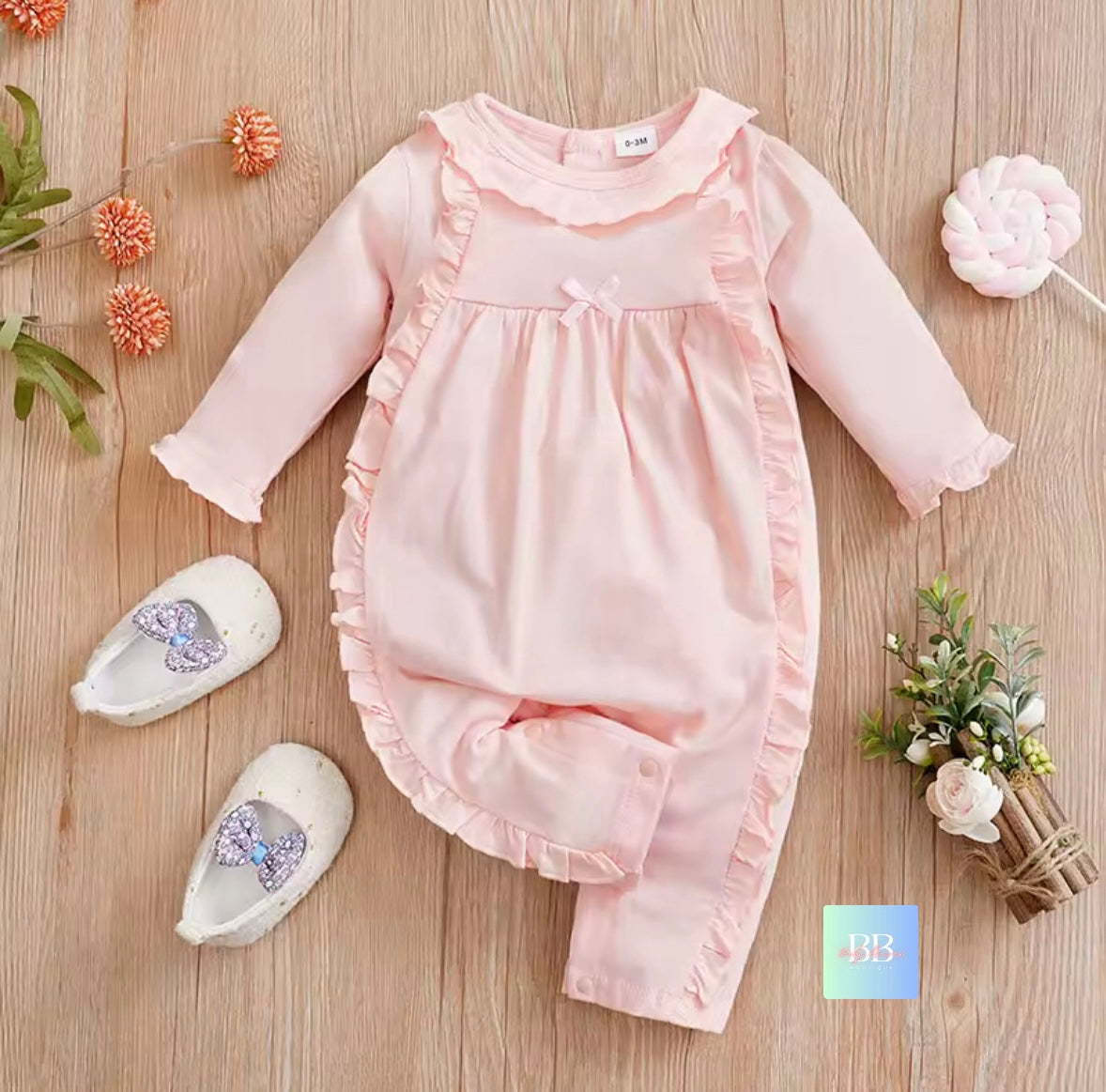 Pink baby romper with ruffles on a wooden surface with decorative items.