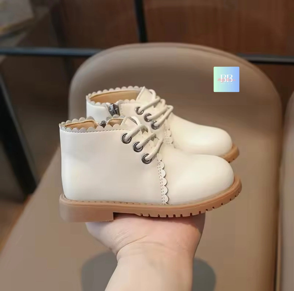 White children's shoes with brown soles held by a hand against a neutral background