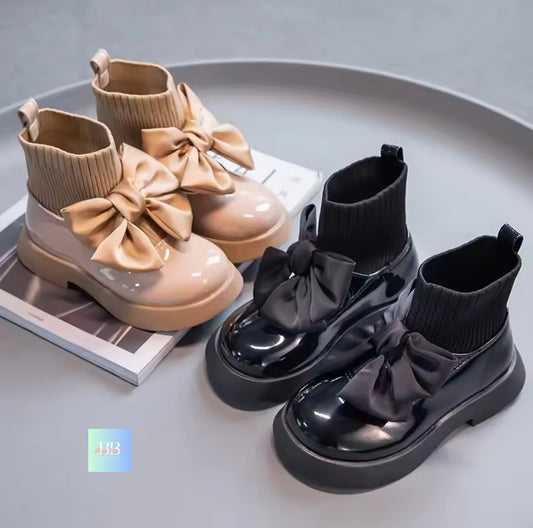 Pair of beige and black shiny boots with large bows on a gray surface.