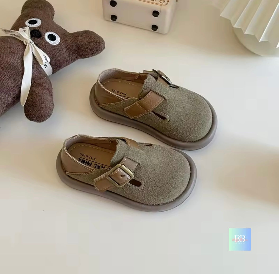 Pair of green children's shoes with buckles on a white surface, next to a small brown teddy bear.