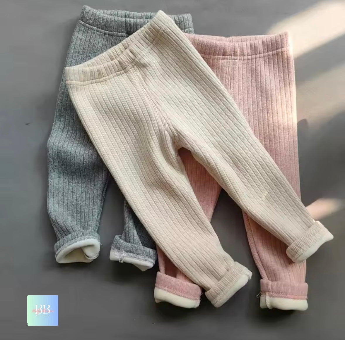 Three pairs of ribbed knit pants in gray, beige, and pink on a dark background.
