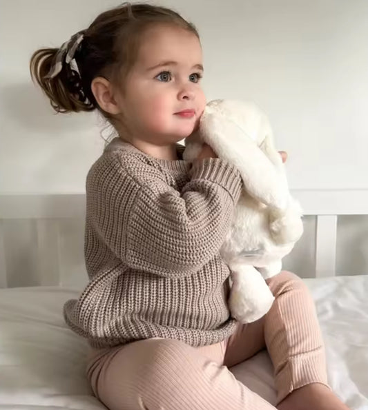 Child wearing a brown knitted sweater and beige pants holding a white fluffy toy.