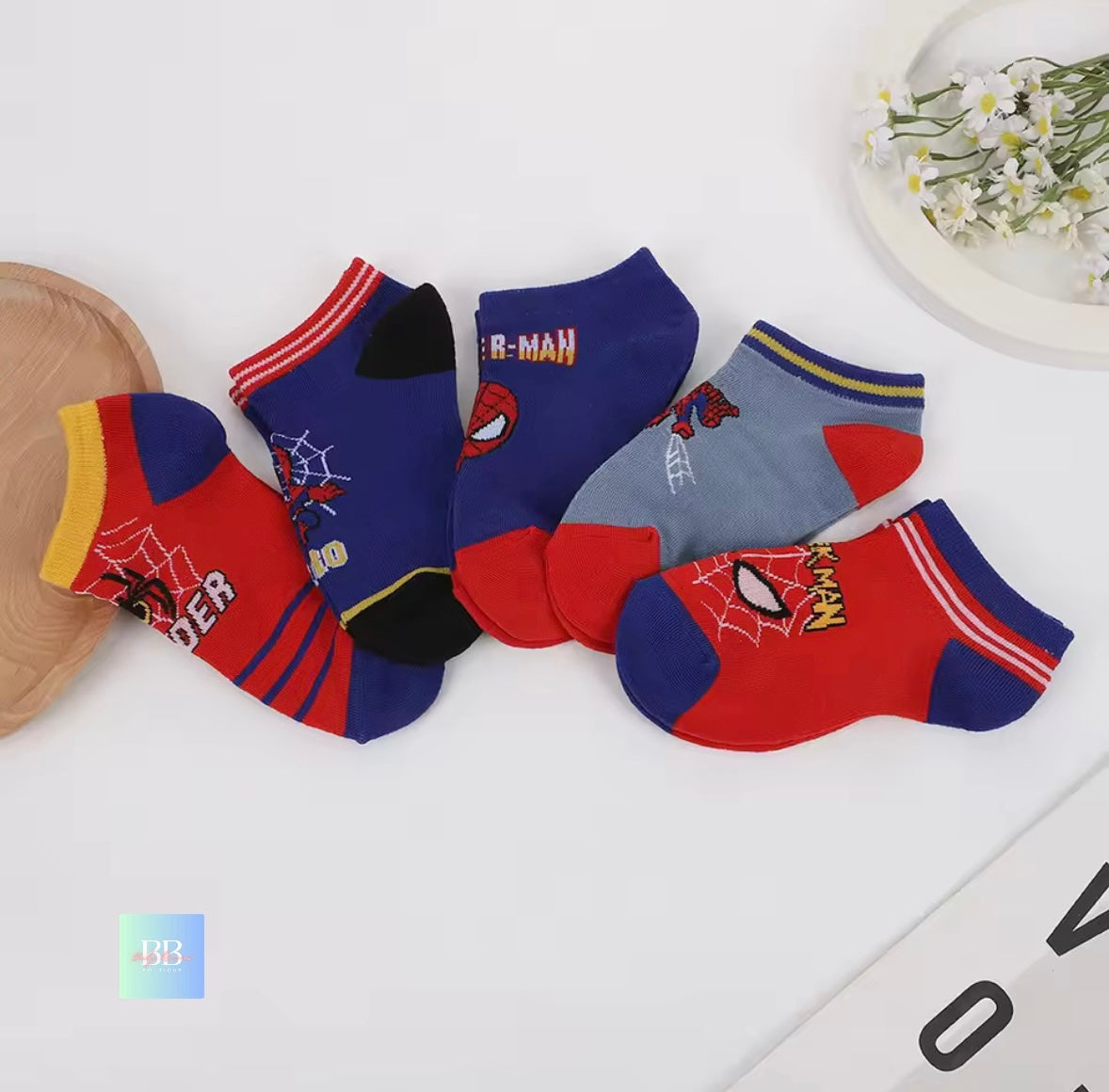 Set of Spider-Man themed socks on a white surface with a decorative plate and flowers in the background.