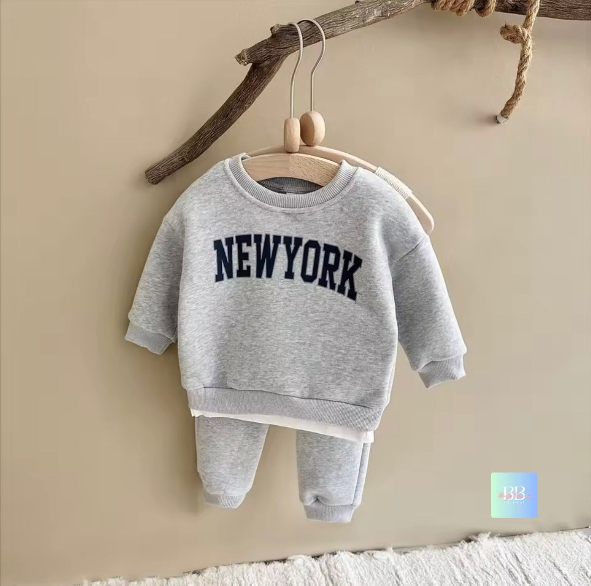 Gray 'New York' sweatshirt and pants set hanging on a wooden hanger against a beige wall.