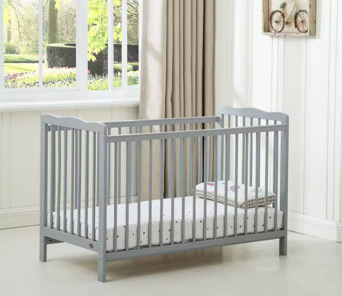 Gray crib with white bedding in a room with a window and curtains.