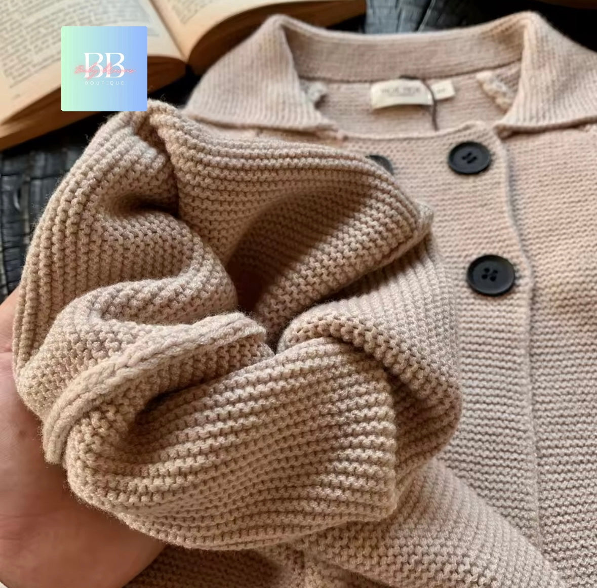 Close-up of a beige knitted sweater with black buttons, held by a hand.