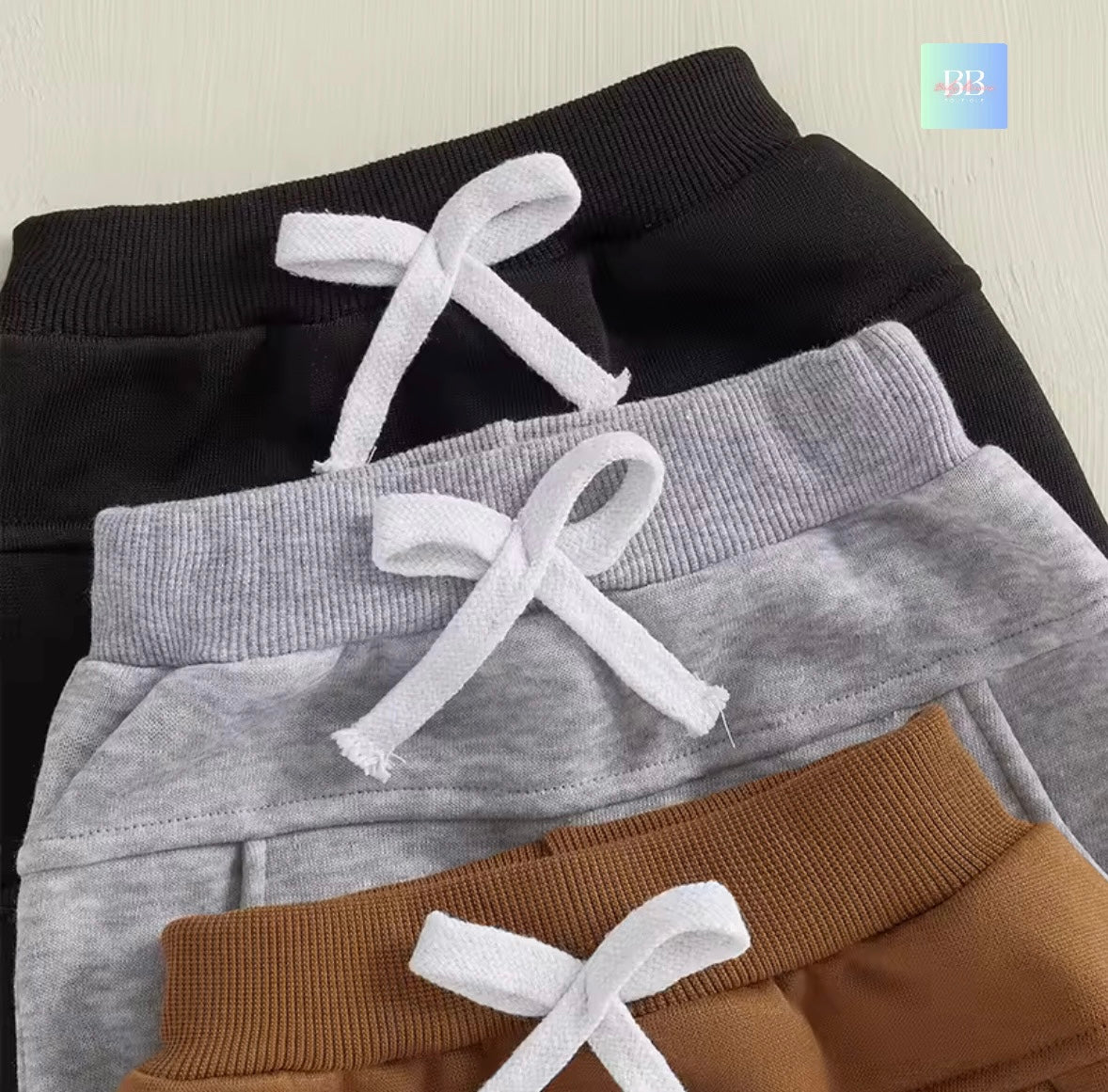 Three pairs of children's jogger pants in black, gray, and brown with white drawstrings on a light background.