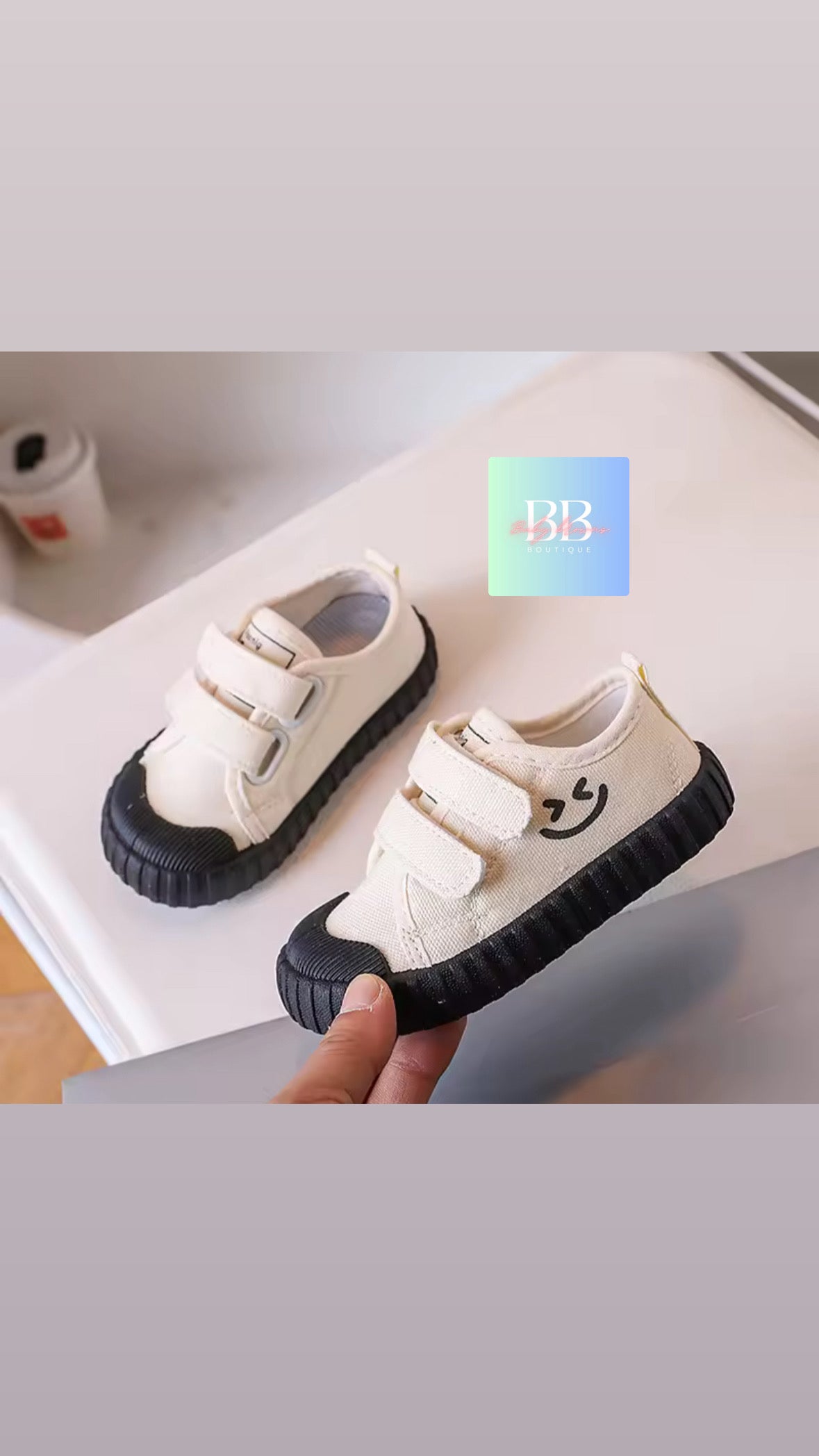 White children's shoes with black soles held by a hand on a light gray surface.